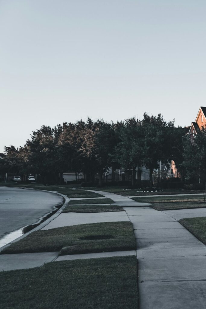 A quiet suburban neighborhood at dusk with empty sidewalks and family homes, suggesting ordinary life continuing after loss